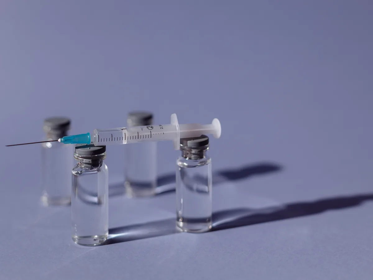 A syringe balanced on vaccine vials with a purple backdrop, representing medical treatment.