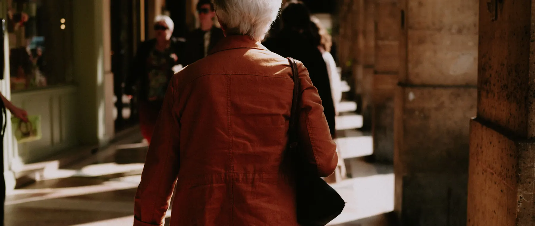 Back view of a senior woman walking along a sunlit city alley, capturing an urban lifestyle moment.