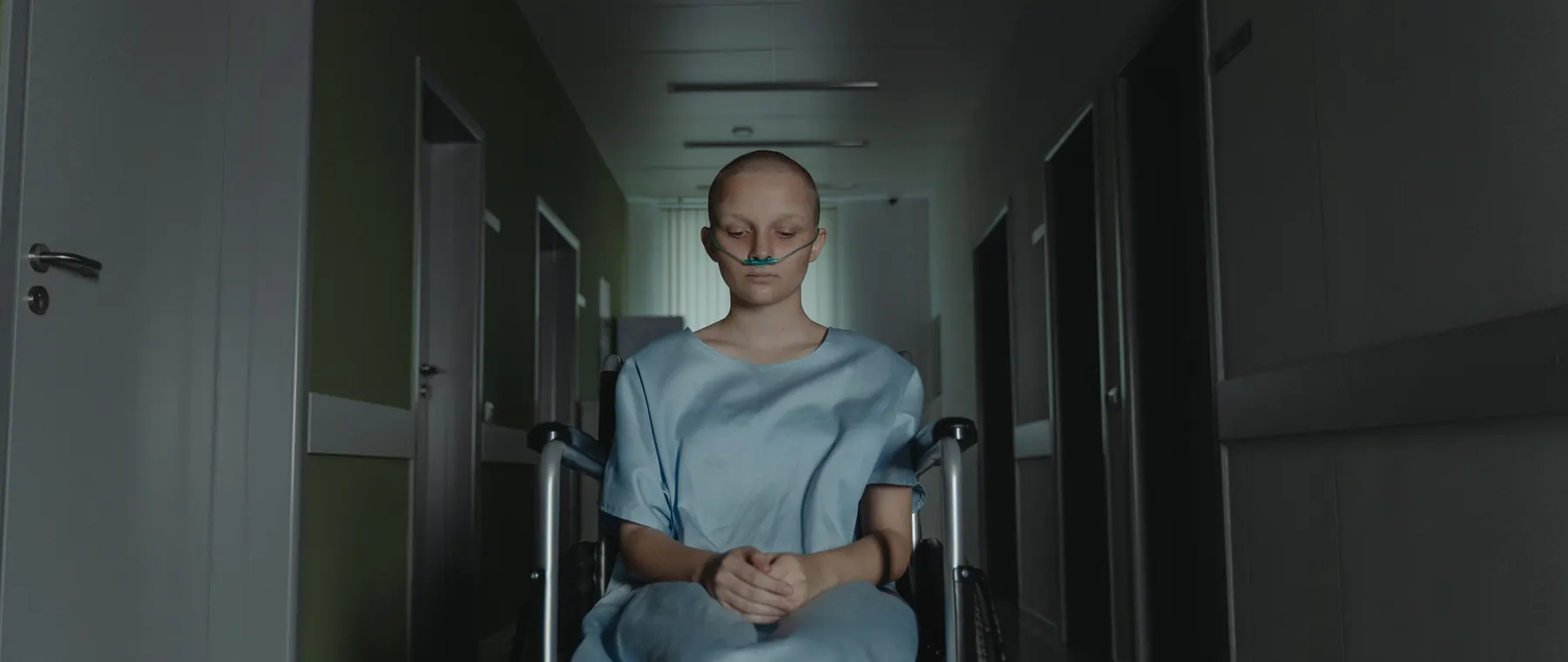 Woman in hospital gown sitting in wheelchair, deep in thought, in dimly lit corridor.