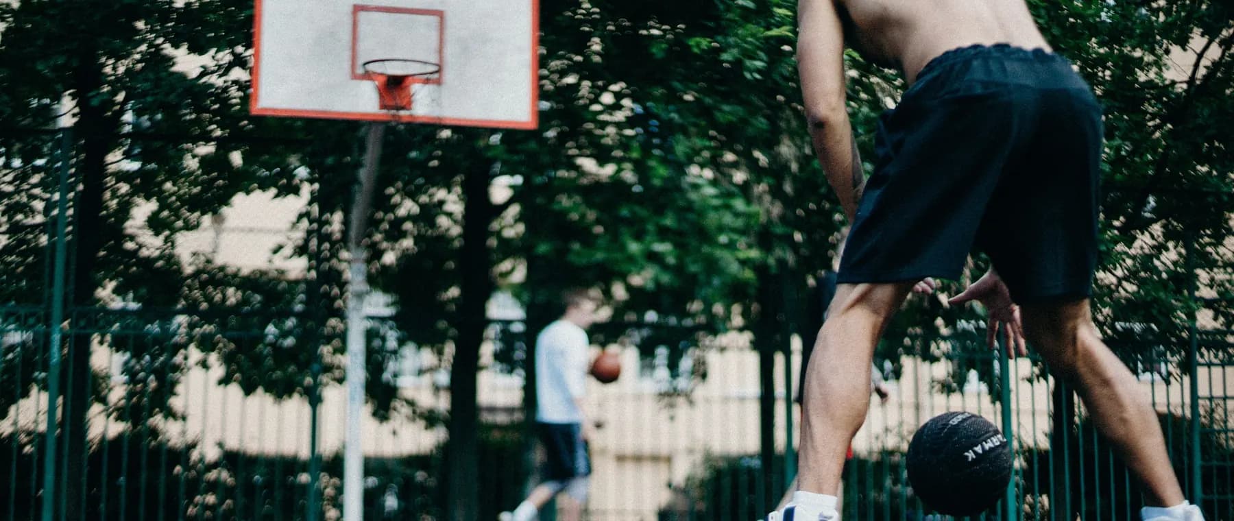 Two athletes playing basketball shirtless on an outdoor court with trees surrounding.