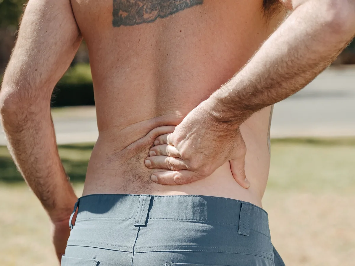 Shirtless man holding his lower back outdoors indicating pain or discomfort.