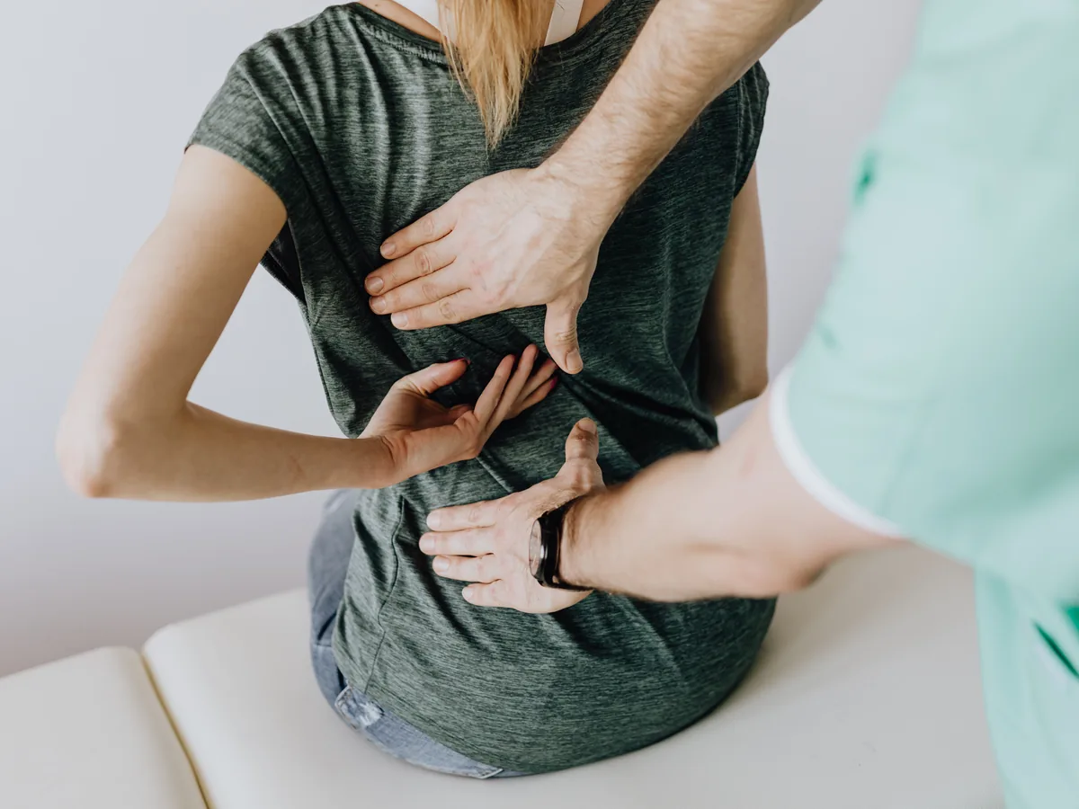 From above of crop faceless slim female patient sitting on examination couch and indicating osteopath in uniform pain spot on back in clinic