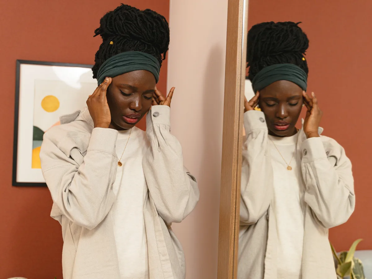 A young woman with braided hair holds her temples in front of a mirror.