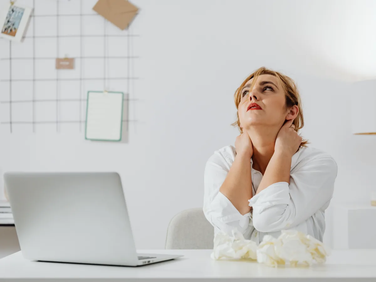 A stressed woman in white long sleeves sits at a desk with a laptop, looking upwards in an office.