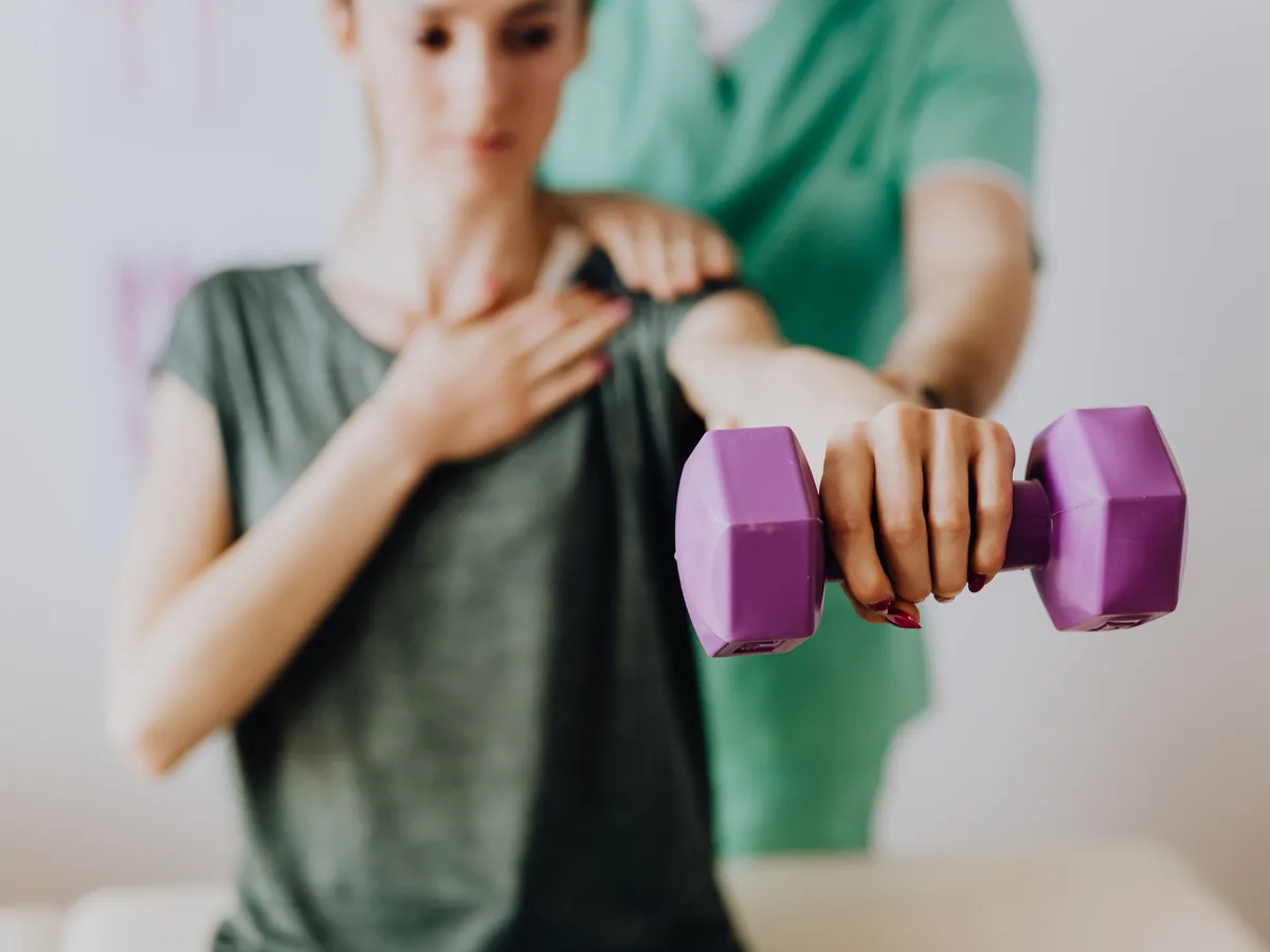 Crop unrecognizable orthopedist in uniform helping slender female in casual wear reaching arm with dumbbell in doctor office in hospital