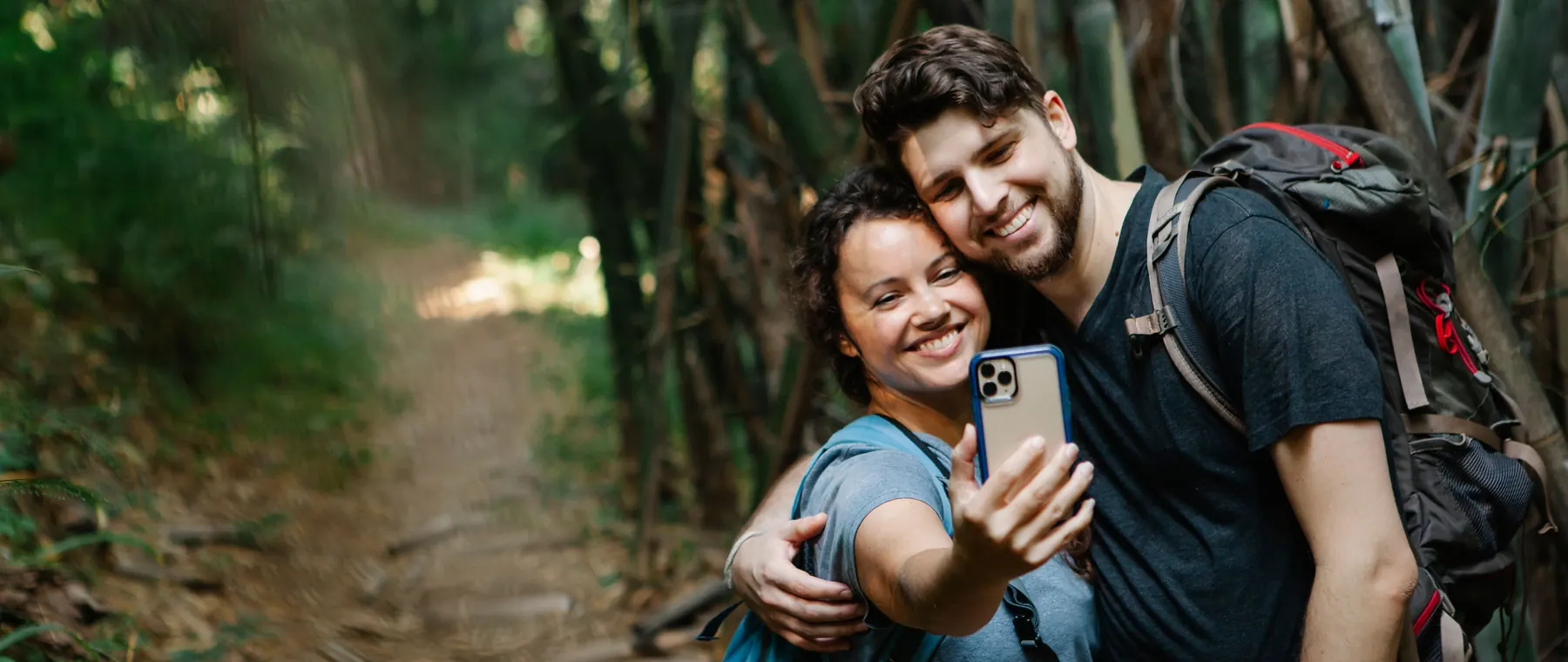 Cheerful young couple in casual clothes and backpacks cuddling and smiling while taking selfie on mobile phone during trekking in green fore