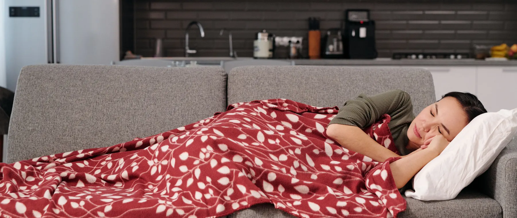 Woman peacefully napping on a couch with a red patterned blanket in a modern kitchen setting.