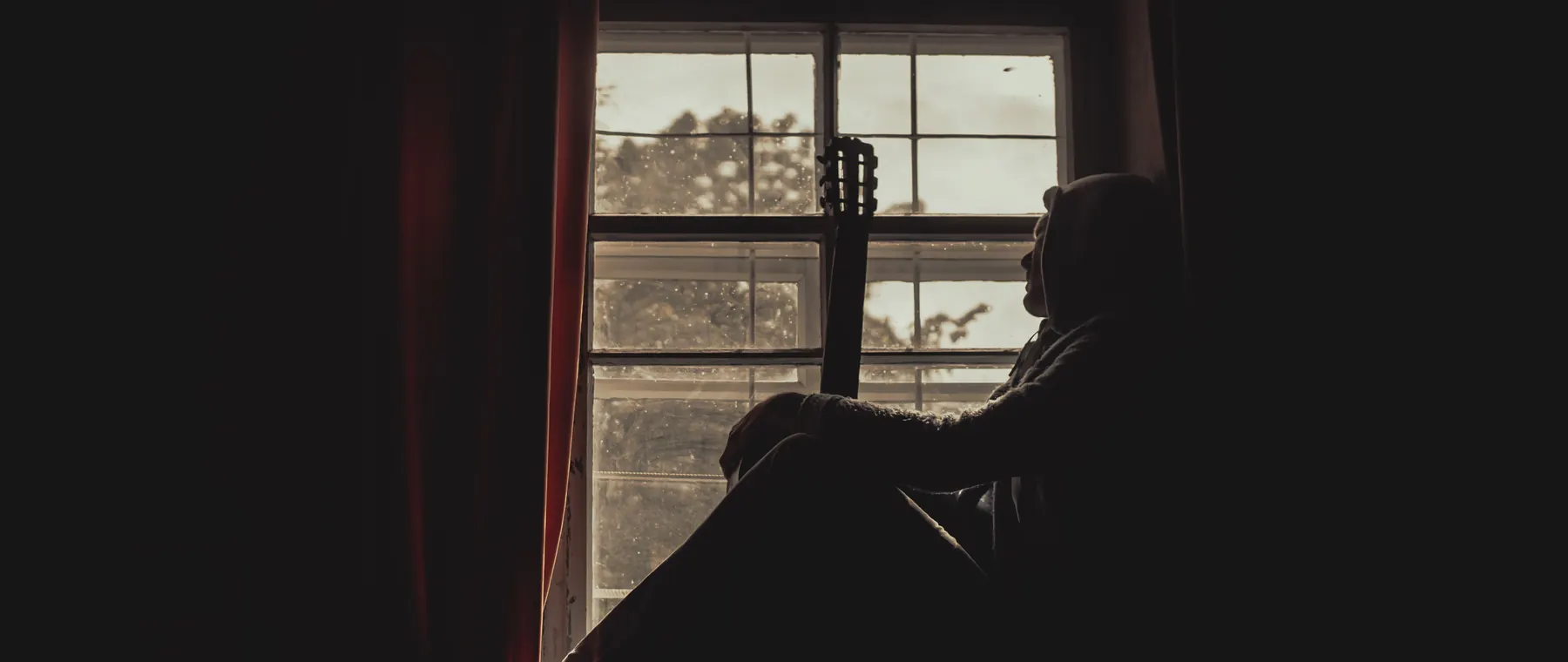 A pensive musician with guitar silhouetted by a window indoors, exuding a moody atmosphere.